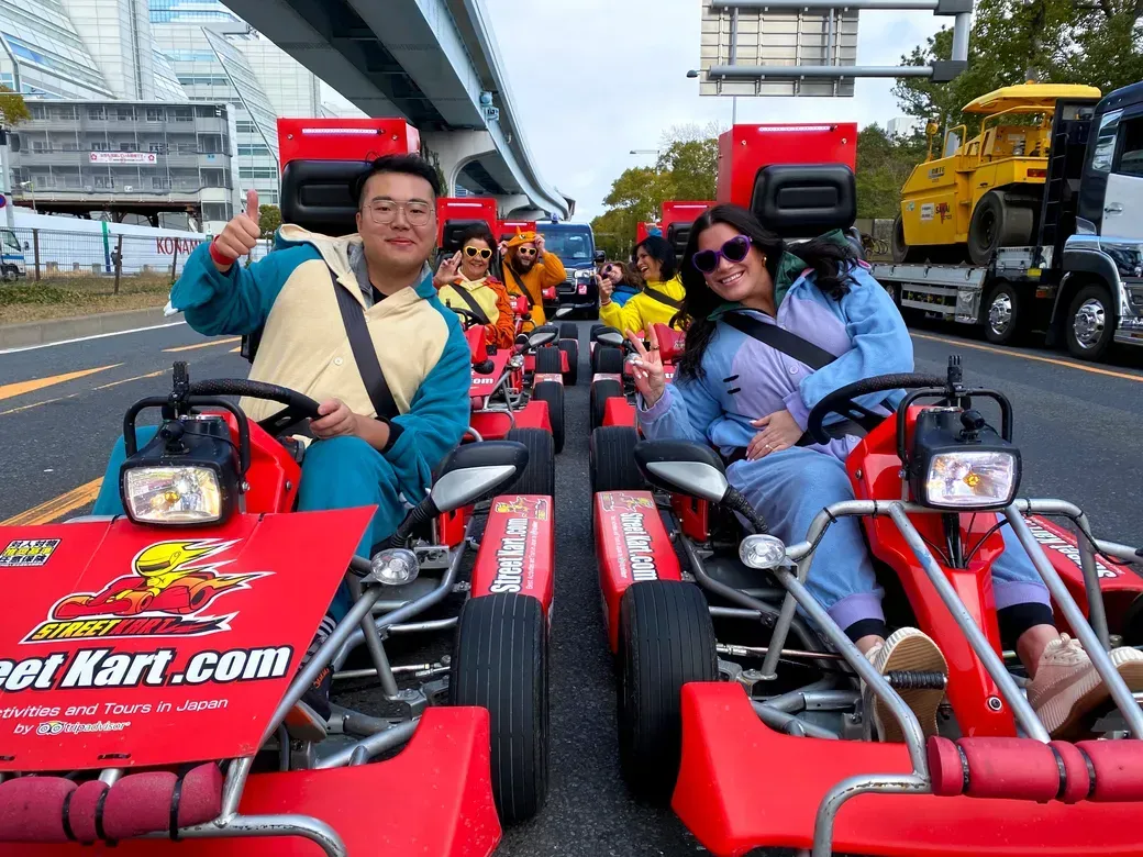 Group of people in red go-karts on a city street, smiling and giving thumbs/peace signs, wearing colorful outfits and sunglasses.