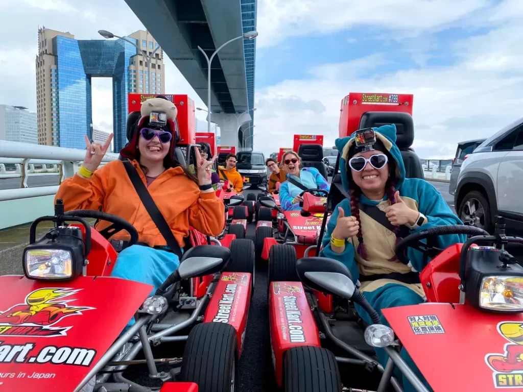 Group of friends in colorful costumes on red go-karts, wearing GoPro head mounts, giving thumbs up on an outdoor track with a city skyline in the background.