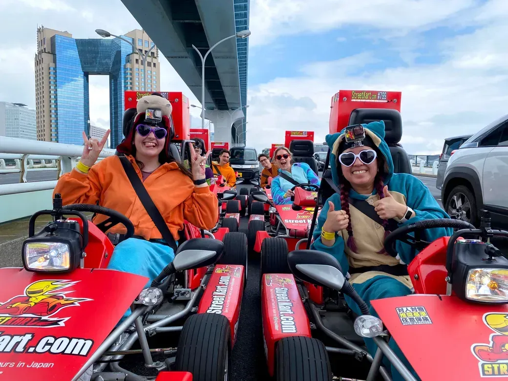 Group of friends in bright costumes sitting in red go-karts on a boardwalk, wearing GoPro hats and posing with smiles and peace signs under a blue sky.