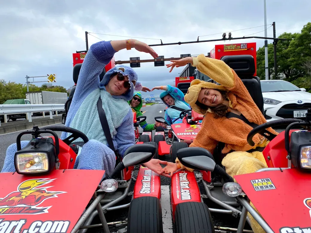 Four people in furry animal costumes posing in red go-karts on a road, forming a heart shape with their arms.