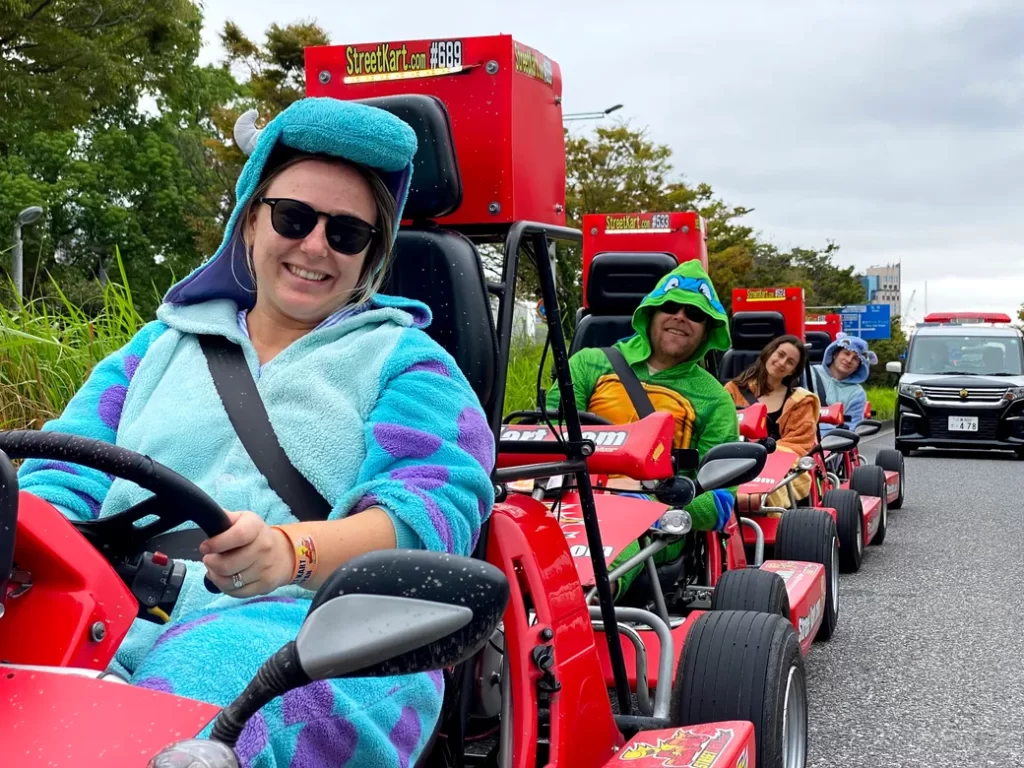 IMG_6175 - StreetKart People in bright costumes ride red go-karts in a parade along a road, smiling at the camera.